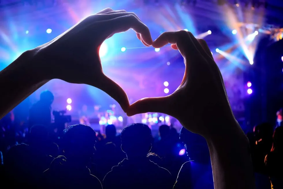 hands forming a heart symbol with a concert scene in the background